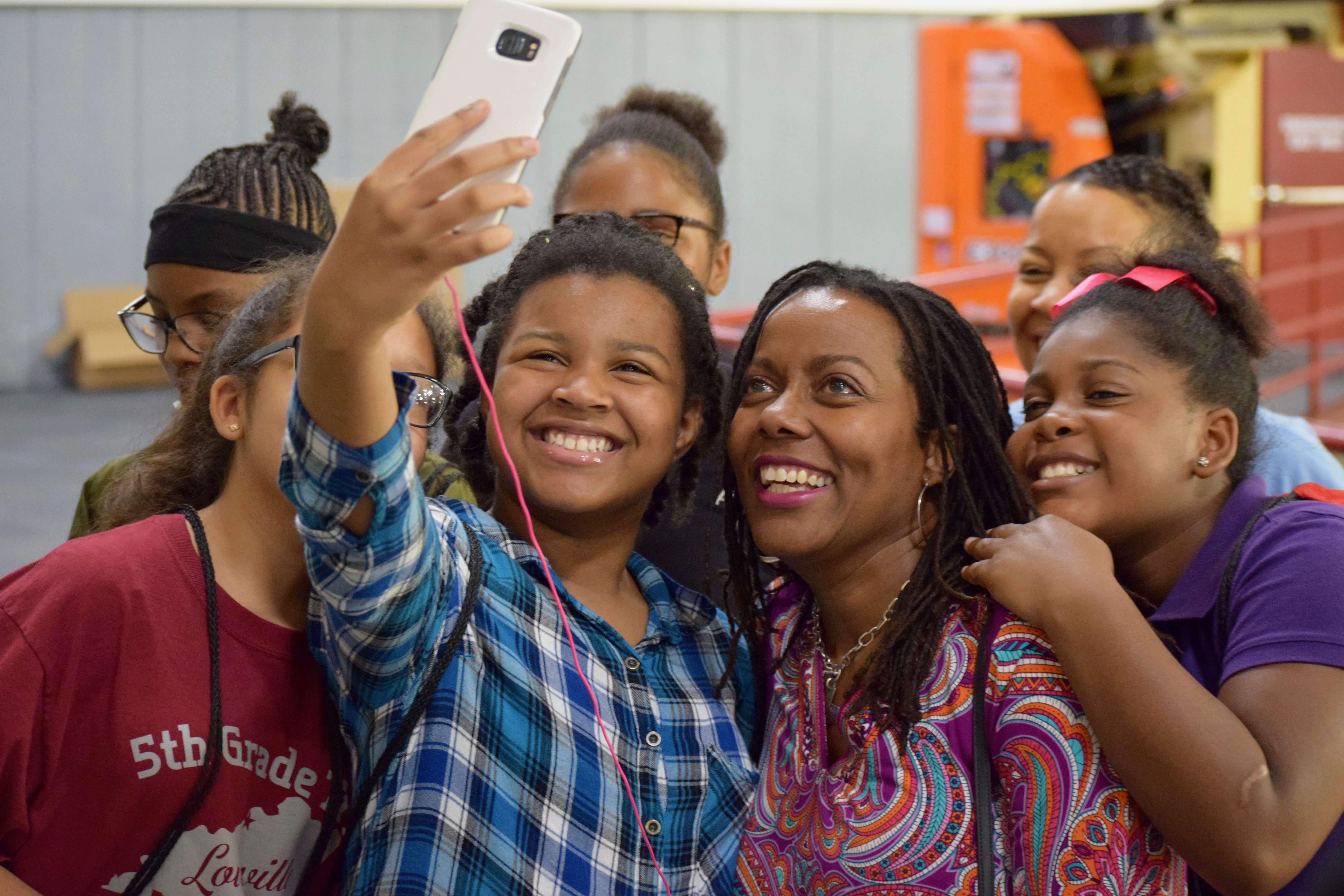 A group of smiling people, including children and an adult, gather closely together indoors as they take a selfie with a smartphone. The atmosphere is cheerful, and everyone is dressed casually.
